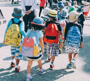 A row of school children going for a walk in the sunshine with colourful backpacks and hats
