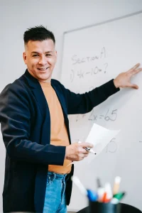 A teacher within a classroom pointing at a whiteboard and smiling